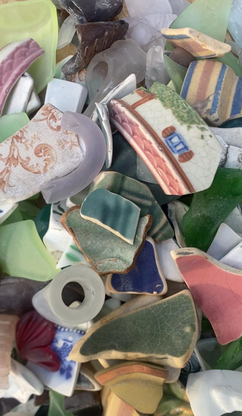 May include: A collection of sea glass and pottery shards in various colors. Pieces include green, blue, pink, and white fragments, some with painted designs. The assortment suggests a craft or decorative theme, perfect for mosaics or jewelry.