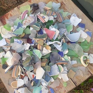May include: A collection of sea glass and pottery shards in various colors, including green, blue, white, and brown, arranged on a weathered wooden surface. The fragments display diverse patterns and textures, suggesting a craft or decorative theme.