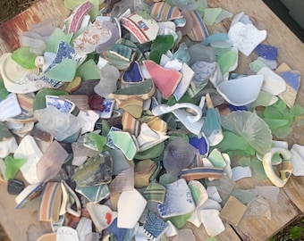 May include: A collection of sea glass and pottery shards in various colors, including green, blue, white, and brown, arranged on a weathered wooden surface. The fragments display diverse patterns and textures, suggesting a craft or decorative theme.