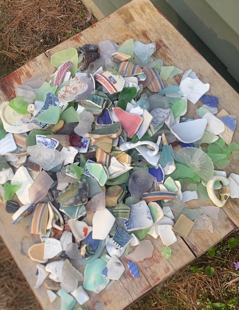 May include: A collection of sea glass and pottery shards in various colors, including green, blue, white, and brown, arranged on a weathered wooden surface. The fragments display diverse patterns and textures, suggesting a craft or decorative theme.