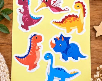 May include: A sheet of six colourful dinosaur stickers on a yellow background. The set includes a purple T-Rex, a pink pterodactyl, a yellow stegosaurus, an orange theropod, a blue triceratops, and a light blue long-necked sauropod dinosaur.