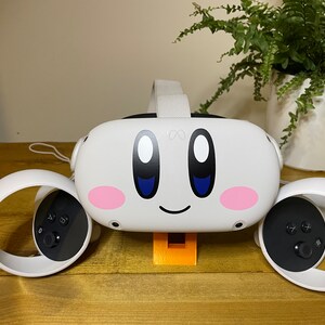 May include: A white virtual reality headset with a pink and blue Kirby character design. The headset is on a stand with two white controllers on either side.