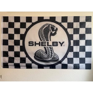 May include: A rectangular flag featuring a black and white checkered pattern on the left and right sides. The center displays a white circle with a black cobra emblem and the word "SHELBY" in bold black letters. The flag is designed for display.