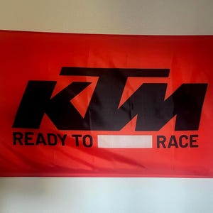 May include: A rectangular red flag with the black KTM logo and the words "READY TO RACE" in black. The flag is hanging on a wall and has grommets for hanging.