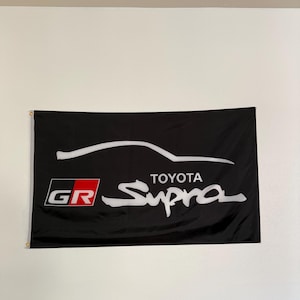 May include: Black flag featuring the Toyota Supra logo in white. The flag includes the GR logo in red and white. A white car outline is above the Toyota Supra text.
