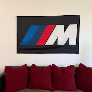 May include: Black flag with the BMW M logo. The logo features three colored stripes in blue, dark blue, and red, with a white "M" on the right. The flag is hanging on a wall above a brown couch with red pillows.