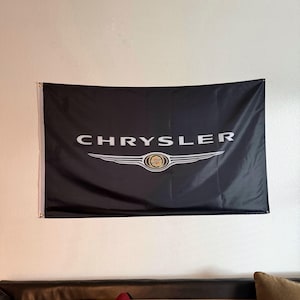 May include: Black flag with the white "CHRYSLER" logo and emblem. The flag is hanging on a white wall above a brown couch with red pillows.