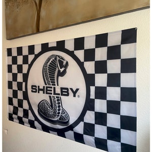 May include: A black and white checkered flag with the word "SHELBY" in bold white letters. The flag features a circular emblem with a cobra snake design. The flag is hanging on a wall.