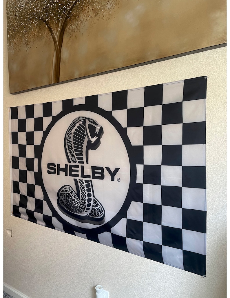 May include: A black and white checkered flag with the word "SHELBY" in bold white letters. The flag features a circular emblem with a cobra snake design. The flag is hanging on a wall.