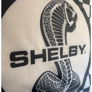 May include: A white flag featuring a large, stylized cobra emblem in black and silver, with the word "SHELBY" in bold black letters. The flag has a checkered border.