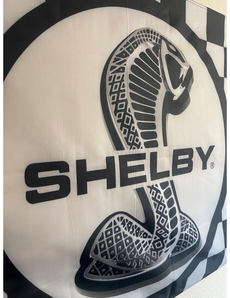 May include: A white flag featuring a large, stylized cobra emblem in black and silver, with the word "SHELBY" in bold black letters. The flag has a checkered border.