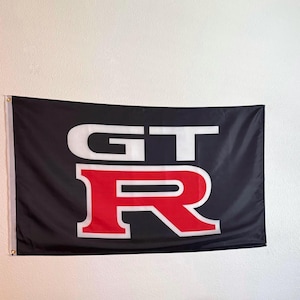 May include: Black flag with the white letters "GT" above a red "R". The flag is rectangular and hangs against a white wall. The flag has a gold-colored grommet on each corner.