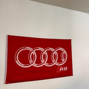 May include: A red flag with the Audi logo in white, featuring four interlocking rings. The letters "RS" are in a white rectangle in the lower right corner. The flag is hanging on a white wall.