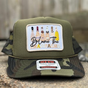 Op de afbeelding: Een camouflage trucker pet met een patch met geïllustreerde wijnflessen en de tekst "Blame The Champagne". De pet heeft een groene kroon, een camo klep en een mesh achterkant.