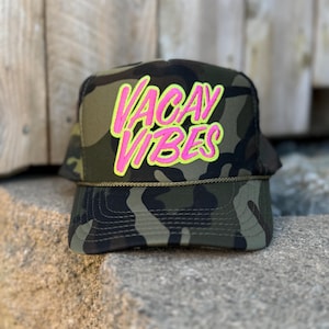 Op de afbeelding: Een camouflage truckerpet met de tekst "VACAY VIBES" in felroze en geel. De pet heeft een gebogen klep en een mesh achterkant, perfect voor een casual, outdoor look.