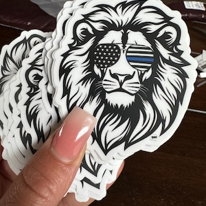 Peut inclure: Une pile d'autocollants représentant une tête de lion en noir et blanc avec un drapeau américain et une ligne bleue sur les lunettes de soleil. Les autocollants ont un logo "BMH" en bas.