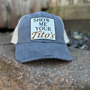 Op de afbeelding: Een distressed blauwe truckerpet met een witte mesh achterkant. De voorkant leest "SHOW ME YOUR Tito's" in witte en oranje letters. De pet ligt op een rots.