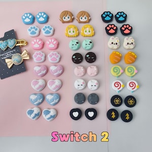 Puede incluir: Una colección de agarres para pulgar Switch 2 coloridos en varios diseños, incluyendo huellas de patas, corazones y caras de personajes. Los agarres son en tonos de azul, rosa, marrón, amarillo, verde, negro, blanco y naranja. El texto "Switch 2" se muestra en rosa en la parte inferior.