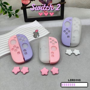 Può includere: Una collezione di accessori per console di gioco Switch 2. L'immagine presenta tre custodie per controller rosa, viola e bianche, insieme a impugnature per pollice a forma di stella. Il testo "Switch 2" è visibile in alto.