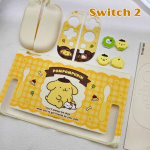 May include: A collection of yellow and white Switch 2 accessories featuring the character Pompompurin. Includes a protective case, controller covers, and thumb grips. The case has a yellow plaid pattern and the text "POMPOMPURIN".