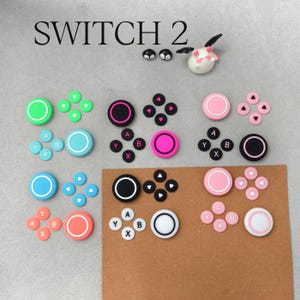 May include: A collection of colorful Switch 2 controller accessories, including thumb grips and button sets in various colors like green, pink, and blue. The image includes the text "SWITCH 2".