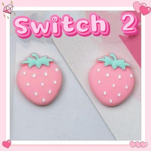 Puede incluir: Dos adornos rosas con forma de fresa con semillas blancas y copas verdes. La palabra "Switch 2" est&aacute; escrita en rosa encima de las fresas. El fondo es un dise&ntilde;o rosa claro y blanco.