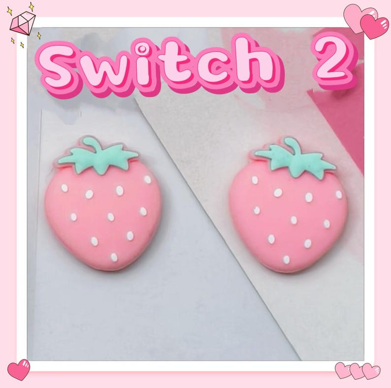 Puede incluir: Dos adornos rosas con forma de fresa con semillas blancas y copas verdes. La palabra "Switch 2" est&aacute; escrita en rosa encima de las fresas. El fondo es un dise&ntilde;o rosa claro y blanco.