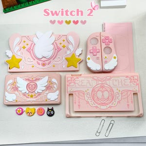 Może przedstawiać: Różowy zestaw akcesoriów do konsoli do gier Switch 2. Zawiera etui na konsolę, osłony kontrolerów i ozdobne zawieszki. Zestaw charakteryzuje się niebiańskim wzorem z gwiazdami, skrzydłami i napisem "DREAMCATCHER".