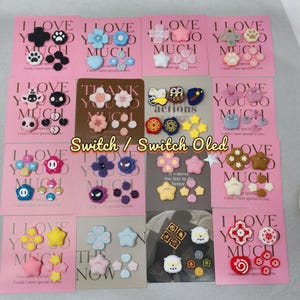以下が含まれることがあります： SwitchまたはSwitch OLED用の装飾アクセサリーのコレクション。アクセサリーには、ピンク、青、白などの色で、花、星、足跡など、さまざまな形が含まれています。各セットは、「I LOVE YOU SO MUCH」というテキストが書かれたピンク色のカードに表示されています。