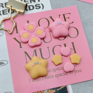 Può includere: Una collezione di abbellimenti a forma di stella e cuore rosa e gialli. Alcuni hanno centri a stella gialli. La frase "I love you so much" è stampata sullo sfondo rosa. Una graffetta dorata è in alto a sinistra.