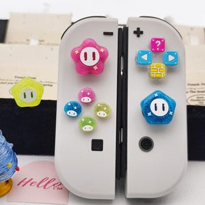 Può includere: Un controller Nintendo Switch bianco con copritasti colorati in stile cartone animato. I copritasti includono una stella rosa, forme di funghi blu e verdi e un pentagono blu. I pulsanti hanno facce bianche e accenti glitterati.