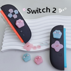 May include: Black and orange Nintendo Switch Joy-Con controllers with colorful, star-shaped button covers. The button covers are pink, blue, and purple with smiling faces. The text "Switch 2" is in the upper right corner.