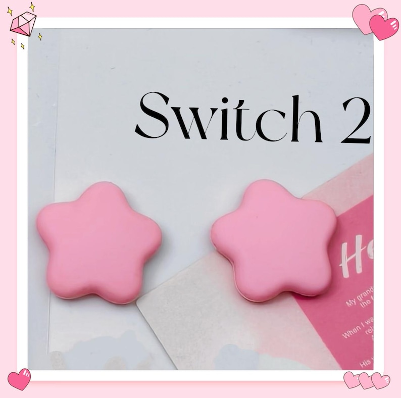 Puede incluir: Dos objetos rosas en forma de estrella. Los objetos son de color rosa suave y tienen un dise&ntilde;o redondeado de cinco puntas. El texto "Switch 2" est&aacute; impreso en negro encima de las estrellas.