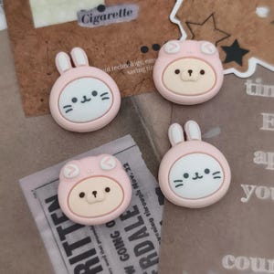 Peut inclure: Quatre breloques roses et blanches représentant des personnages de dessins animés. Deux breloques présentent un motif de lapin et deux un motif d'ours. Les breloques sont rondes et ont un visage blanc avec des détails noirs. Le mot "Cigarette" est visible en arrière-plan.