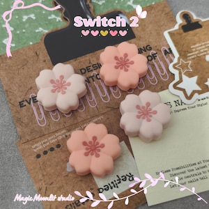 Könnte beinhalten: Vier blütenförmige Nagelkunstdekorationen in Rosa- und Pfirsichtönen. Die Dekorationen sind auf einem braunen Papierhintergrund mit dem Text "Switch 2" und "Magic Moonlit studio" angeordnet.