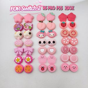 Pode incluir: Uma coleção de capas de joystick rosa com vários designs, incluindo estrelas, morangos, pêssegos, laços e rostos de personagens. O texto "FOR Switch 2 NS PRO PS5 XBOX" está visível no topo.