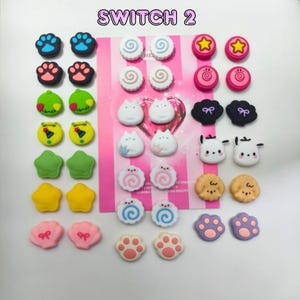 Puede incluir: Una colección de botones decorativos coloridos con varios diseños, incluyendo huellas de patas, estrellas y caras de animales. El texto "SWITCH 2" se muestra en la parte superior. Los botones están dispuestos sobre un fondo rosa.