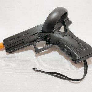 Puede incluir: Controlador de pistola VR negro con punta naranja. El controlador tiene el texto "Q1VR 17 IRELAND" impreso en el lateral. Una correa negra está unida a la parte inferior del controlador.