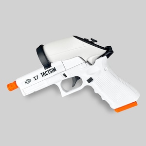 Puede incluir: Pistola de entrenamiento K3D 17 TACTSIM blanca y negra con punta y base naranja. La pistola tiene una empuñadura blanca y una correa negra que sujeta una pieza superior blanca. El texto "K3D 17 TACTSIM" está impreso en el lateral.