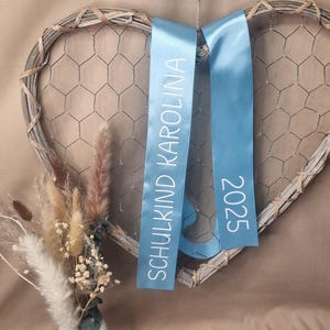 May include: A heart-shaped wall decoration made of grey woven wood and wire mesh, featuring two light blue satin ribbons hanging down. The left ribbon displays the white text SCHULKIND KAROLINA, and the right ribbon displays the white text 2025.
