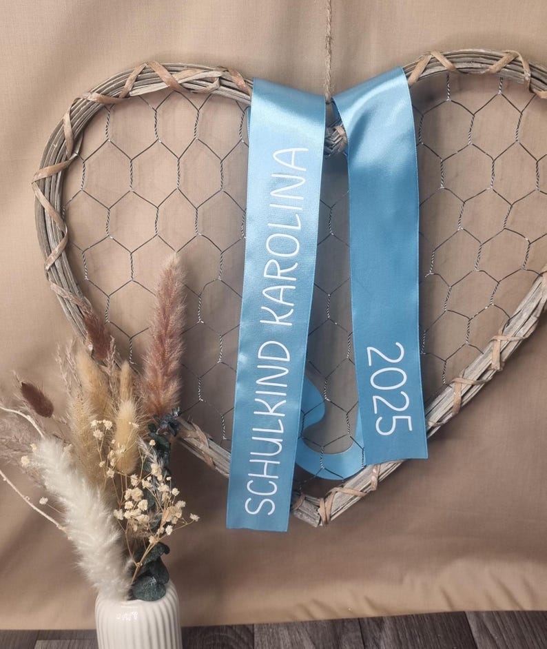 May include: A heart-shaped wall decoration made of grey woven wood and wire mesh, featuring two light blue satin ribbons hanging down. The left ribbon displays the white text SCHULKIND KAROLINA, and the right ribbon displays the white text 2025.