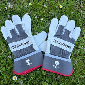 May include: A pair of work gloves with the text "CHEF HANDWERKER" printed on the wristband. The gloves are light blue and gray with red trim and the "STRAUSS 10" logo. They are lying on a bed of green grass.