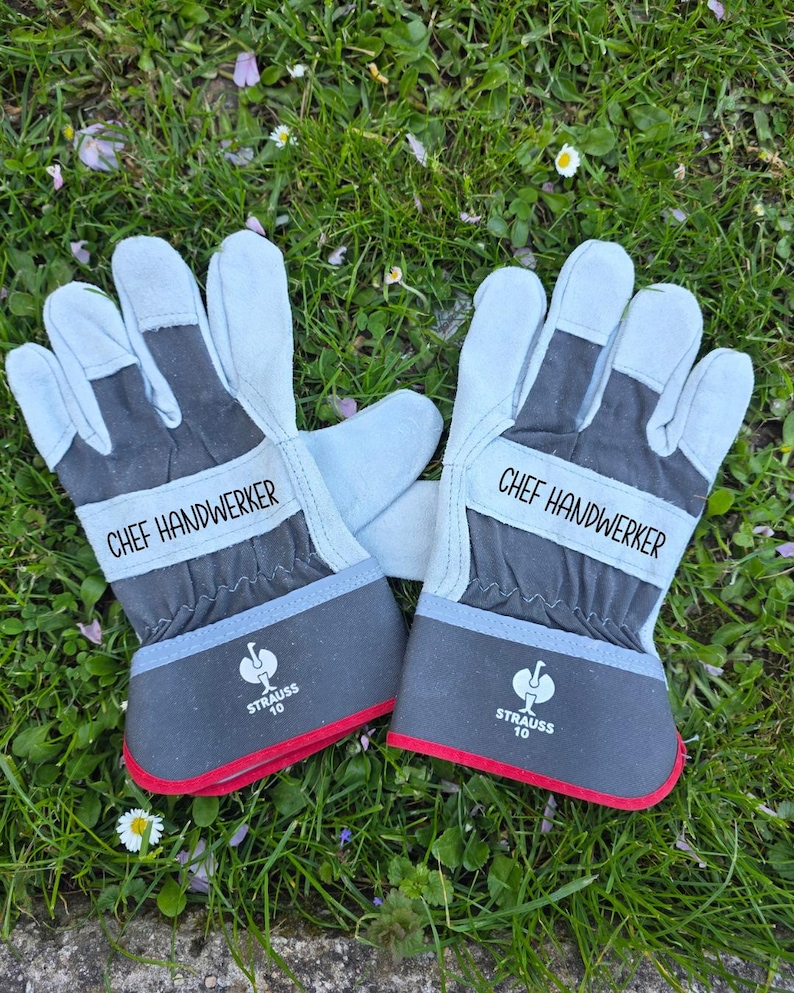 May include: A pair of work gloves with the text "CHEF HANDWERKER" printed on the wristband. The gloves are light blue and gray with red trim and the "STRAUSS 10" logo. They are lying on a bed of green grass.