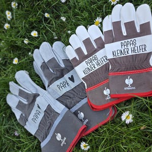 May include: A set of work gloves with white, gray, and brown accents, resting on a bed of green grass and daisies. The gloves feature the word "PAPA" and "PAPAS KLEINER HELFER" printed on them, along with a heart symbol.