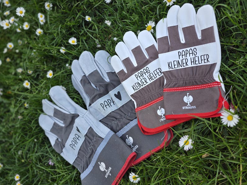 May include: A set of work gloves with white, gray, and brown accents, resting on a bed of green grass and daisies. The gloves feature the word "PAPA" and "PAPAS KLEINER HELFER" printed on them, along with a heart symbol.
