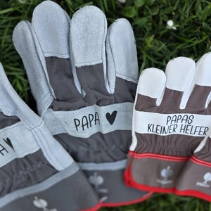 May include: Gray and brown work gloves with white fingertips. The gloves have the word "PAPA" printed on them with a heart symbol. Another pair of gloves has the text "PAPAS KLEINER HELFER" printed on them. Red trim accents the gloves.