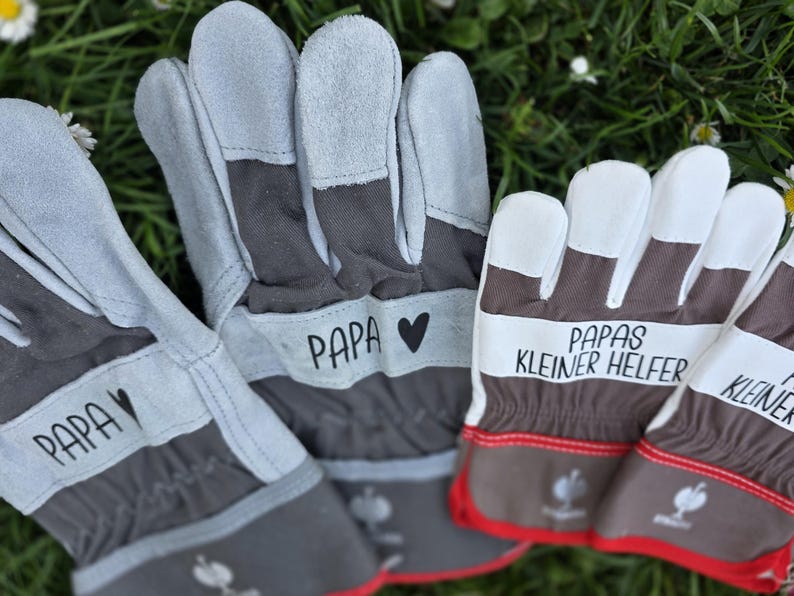 May include: Gray and brown work gloves with white fingertips. The gloves have the word "PAPA" printed on them with a heart symbol. Another pair of gloves has the text "PAPAS KLEINER HELFER" printed on them. Red trim accents the gloves.