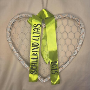 May include: A heart-shaped wall decoration featuring a light-colored woven frame filled with silver wire mesh. Two lime green satin ribbons hang from the top, printed with the black text SCHULKIND ELIAS on one side and the year 2025 on the other.