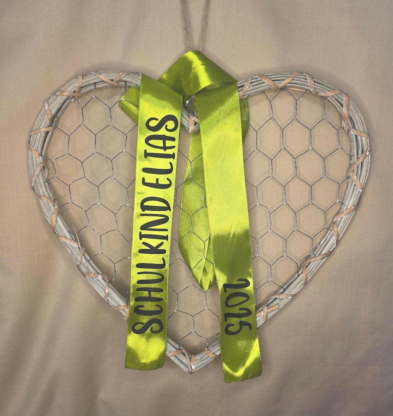 May include: A heart-shaped wall decoration featuring a light-colored woven frame filled with silver wire mesh. Two lime green satin ribbons hang from the top, printed with the black text SCHULKIND ELIAS on one side and the year 2025 on the other.