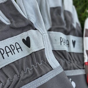 May include: Close-up of gray and light gray gardening gloves. The gloves have the word "PAPA" printed on a light gray band, along with a black heart. The gloves are made of a durable material and are designed for gardening.